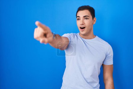 Photo for Young hispanic man standing over blue background pointing with finger surprised ahead, open mouth amazed expression, something on the front - Royalty Free Image