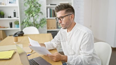 Photo for Handsome young hispanic business professional fully immersed in reading critical documents at office desk, looking serious and concentrated - Royalty Free Image