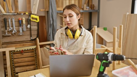 Caucasian brunette woman using smartphone in a carpentry workshop with tools and woodwork.