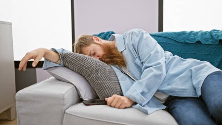 Erschöpfter junger irischer Mann mit rothaarigem Bart entspannt sich auf gemütlichem Sofa, schläft tief zu Hause im Wohnzimmer und genießt die Entspannung einer wohlverdienten Erholung