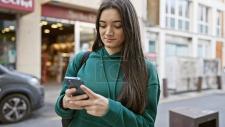 Foto de Una joven hispana usa su smartphone en una bulliciosa calle de la ciudad, personificando la vida urbana y la tecnología. - Imagen libre de derechos