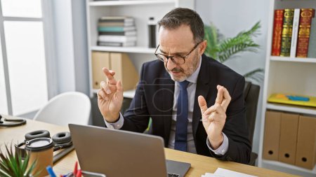 Téléchargez les photos : Coup de chance pour un homme sérieux d'âge moyen barbu aux cheveux gris, travaillant en ligne sur un ordinateur portable à son bureau, croisant les doigts pour réussir - en image libre de droit