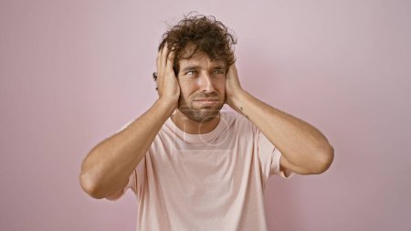 Foto de Un joven hispano preocupado con una camisa rosa se levanta contra una pared rosa, mirando frustrado con las manos en la cabeza. - Imagen libre de derechos