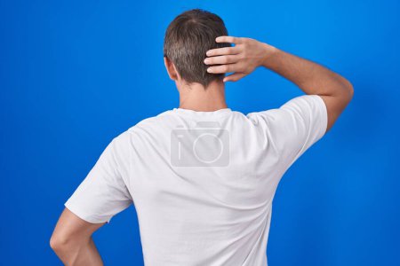 Photo for Caucasian blond man standing over blue background backwards thinking about doubt with hand on head - Royalty Free Image
