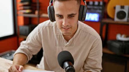 A young adult hispanic man in a music studio, wearing headphones and engaging with a microphone.