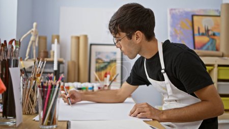 Téléchargez les photos : Talentueux jeune artiste hispanique entièrement absorbé par le dessin au crayon dans un atelier d'art animé, mettant en valeur le talent brut au milieu des pinceaux et de la toile - en image libre de droit
