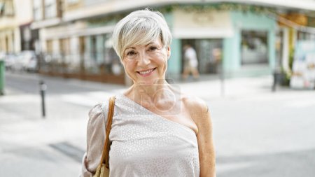 Mature hispanic woman with short grey hair smiling on a sunny urban street.