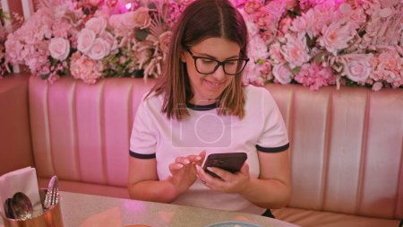 Foto de Mujer sonriente usando smartphone en un café rosa adornado con flores - Imagen libre de derechos