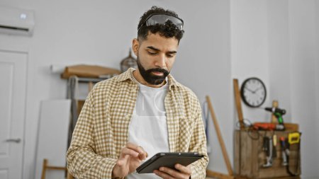 Photo for A handsome young hispanic man with a beard works on a tablet in a well-organized carpentry workshop. - Royalty Free Image