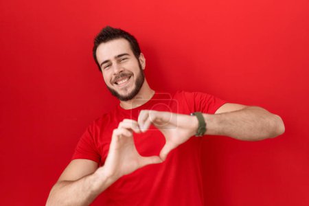 Téléchargez les photos : Jeune homme hispanique portant un t-shirt rouge décontracté souriant en amour faisant forme de symbole de coeur avec les mains. concept romantique. - en image libre de droit