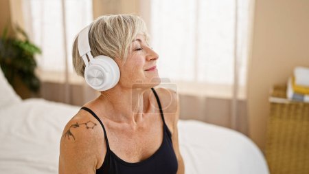 Téléchargez les photos : Une femme mûre détendue aux cheveux gris profitant de la musique sur écouteurs dans un cadre de chambre confortable. - en image libre de droit