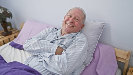 Téléchargez les photos : Joyeux homme âgé, aux cheveux gris et confiant, couché dans une chambre confortable avec les bras croisés, profitant tranquillement d'une matinée détendue en pyjama - en image libre de droit