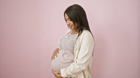 Cheery young pregnant woman with a radiant smile relishes joyfully touching her belly, standing casually over pink isolated background