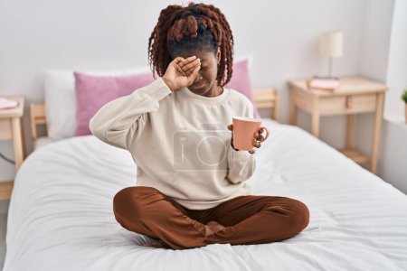 Foto de Mujer afroamericana sentada en la cama bostezando y tomando café en el dormitorio - Imagen libre de derechos