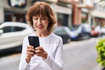 Téléchargez les photos : Femme d'âge moyen souriant confiant en utilisant smartphone à la rue - en image libre de droit