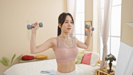 Photo for A young asian woman exercises with dumbbells in a bright, cozy bedroom, embodying fitness and a healthy lifestyle at home. - Royalty Free Image