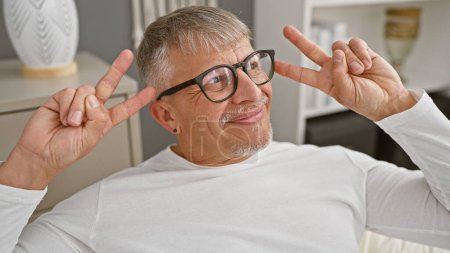 Lächelnder Mann mittleren Alters mit Brille macht Friedenszeichen, während er sich in einem weißen Schlafzimmer entspannt