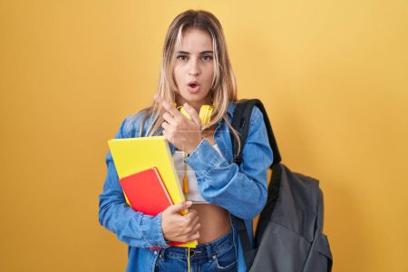 Foto de Mujer rubia joven usando mochila de estudiante y sosteniendo libros sorprendidos señalando con el dedo hacia un lado, boca abierta expresión asombrada. - Imagen libre de derechos