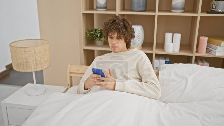 Photo for A young man with curly hair using a smartphone while relaxing in a cozy bedroom setting - Royalty Free Image