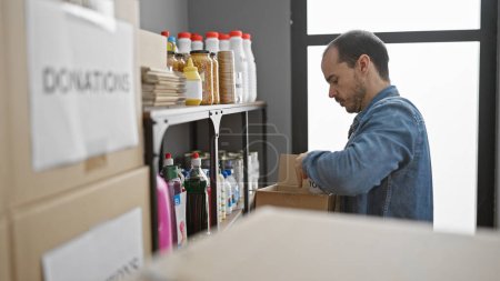 Hombre hispano organizando donaciones en un almacén con estantes de alimentos y suministros en el fondo