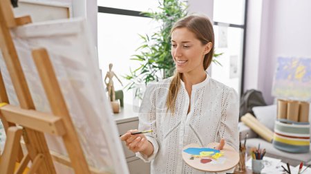 Photo for Smiling caucasian woman painting on canvas in a bright art studio - Royalty Free Image