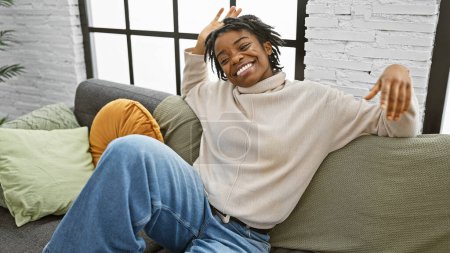 Foto de Mujer afroamericana con rastas sonriendo en un sofá en un acogedor salón interior. - Imagen libre de derechos