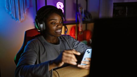 Téléchargez les photos : Jeune femme noire avec dreadlocks portant un casque et utilisant un smartphone dans une salle de jeux faiblement éclairée. - en image libre de droit