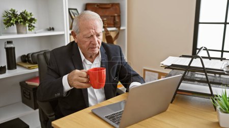 Foto de Elegante hombre mayor, centrado en el éxito, trabajando en línea con el ordenador portátil en el escritorio de la oficina, beber café de la mañana de una manera relajada pero seria. - Imagen libre de derechos