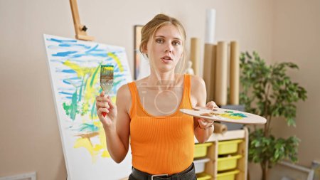 Photo for Young blonde woman artist in studio with paintbrush and palette creating abstract painting on canvas - Royalty Free Image