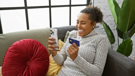 Foto de Mujer sonriente sosteniendo tarjeta de crédito y teléfono inteligente sentado en el sofá en el interior con plantas en el fondo. - Imagen libre de derechos