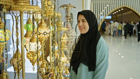 Photo for Smiling woman in hijab admiring traditional lamps at a cultural marketplace in abu dhabi. - Royalty Free Image