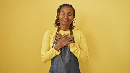 Téléchargez les photos : Femme mûre afro-américaine souriante avec les yeux fermés, main sur le cœur, debout sur fond jaune - en image libre de droit