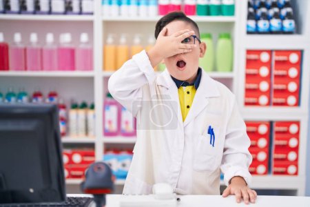 Téléchargez les photos : Jeune enfant hispanique travaillant à la pharmacie drugstore jetant un coup d'oeil dans le visage de couverture de choc et les yeux avec la main, regardant à travers les doigts peur - en image libre de droit