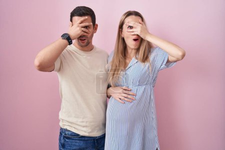 Photo for Young couple expecting a baby standing over pink background peeking in shock covering face and eyes with hand, looking through fingers with embarrassed expression. - Royalty Free Image