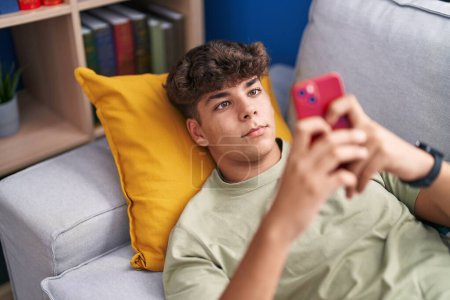 Téléchargez les photos : Jeune adolescent hispanique utilisant un smartphone couché sur le canapé à la maison - en image libre de droit