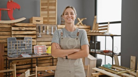 Photo for Portrait of a confident young woman in a workshop, wearing an apron and standing arms crossed amidst woodworking tools. - Royalty Free Image