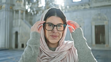 Foto de Una joven sonriente con gafas y un pañuelo en la cabeza posa dentro de una mezquita de Estambul iluminada por el sol, representando la belleza y la diversidad cultural. - Imagen libre de derechos
