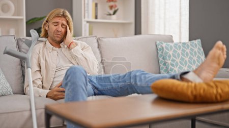 Téléchargez les photos : Un homme qui a mal à la tête s'incline sur un canapé près de béquilles dans un salon bien éclairé. - en image libre de droit