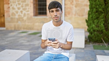 Junger hispanischer Mann benutzt Smartphone mit ernster Miene im Park