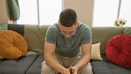 Photo for Pensive young man in glasses sitting on couch indoors, reflecting alone at home. - Royalty Free Image