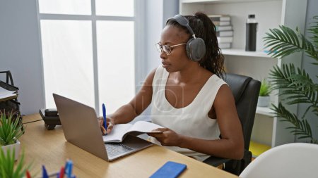 Photo for African american woman wearing headphones taking notes at her office desk indoors - Royalty Free Image