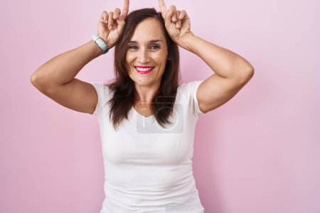 Photo for Middle age brunette woman standing over pink background doing funny gesture with finger over head as bull horns - Royalty Free Image