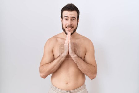 Téléchargez les photos : Jeune homme hispanique debout torse nu sur fond blanc priant avec les mains ensemble demandant pardon en souriant confiant. - en image libre de droit
