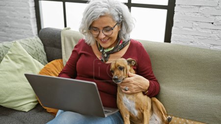 Foto de Una mujer sonriente con el pelo gris acaricia a su perro mientras usa un ordenador portátil en un acogedor interior de la sala de estar. - Imagen libre de derechos
