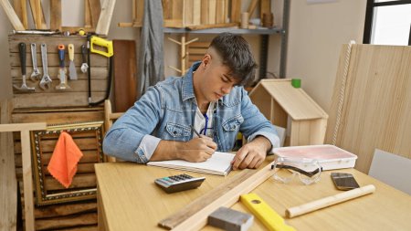 Photo for Attractive young hispanic man, a professional carpenter, engrossed in drawing on a table indoors; the real portrait of a hardworking handyman in his woodworking studio. - Royalty Free Image
