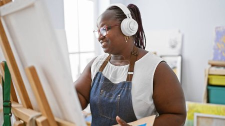 Téléchargez les photos : Artiste afro-américaine confiante dessinant avec passion, écoutant de la musique dans un studio d'art, embrassant ses courbes et souriant - en image libre de droit