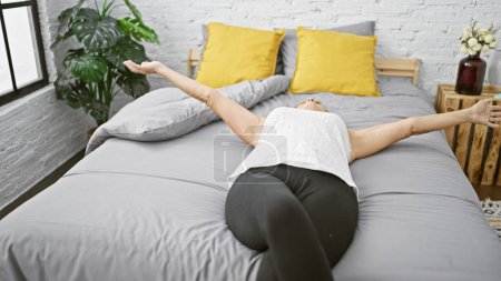 Schöne blonde Frau mittleren Alters kuschelt sich in den Komfort ihres Schlafzimmers, breitet ihre Arme gemächlich aus, während sie auf einem bequemen Bett liegt