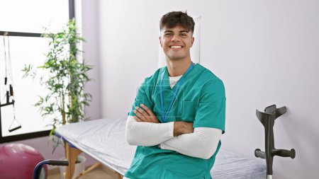 Téléchargez les photos : Jeune homme hispanique confiant et heureux travaillant comme physiothérapeute, souriant avec un geste détendu des bras croisés dans une clinique de réadaptation dynamique. - en image libre de droit