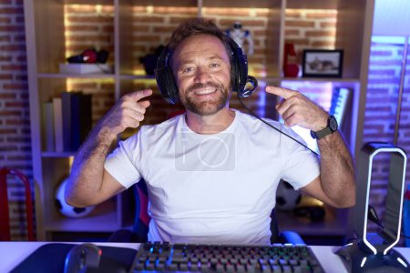 Photo for Middle age man with beard playing video games wearing headphones smiling cheerful showing and pointing with fingers teeth and mouth. dental health concept. - Royalty Free Image