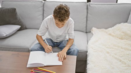 Photo for Cute, adorable little blond boy sitting on sofa at home, drawing intently in notebook, completely lost in the world of his imagination - Royalty Free Image